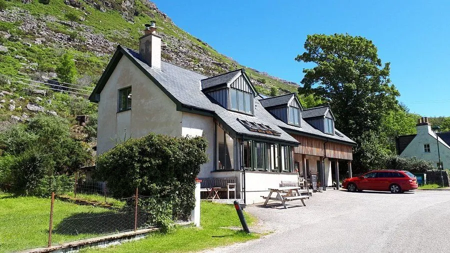 An exterior view of a building nestled against a green mountain under a clear blue sky. The building features multiple windows and dormers, with a small outdoor seating area.
