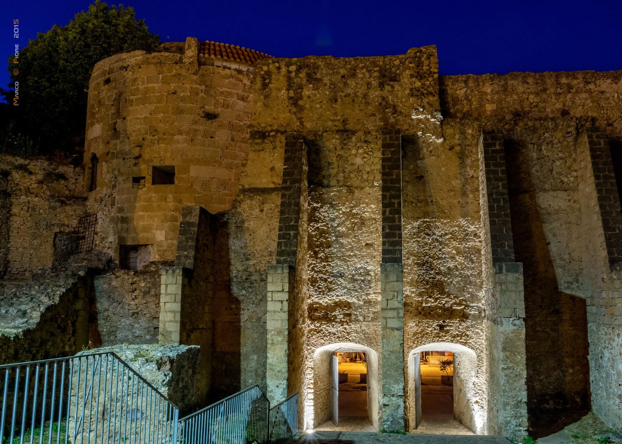 A night view of an ancient stone building, possibly a fortress or castle, featuring illuminated archways at its base under a dark blue sky.