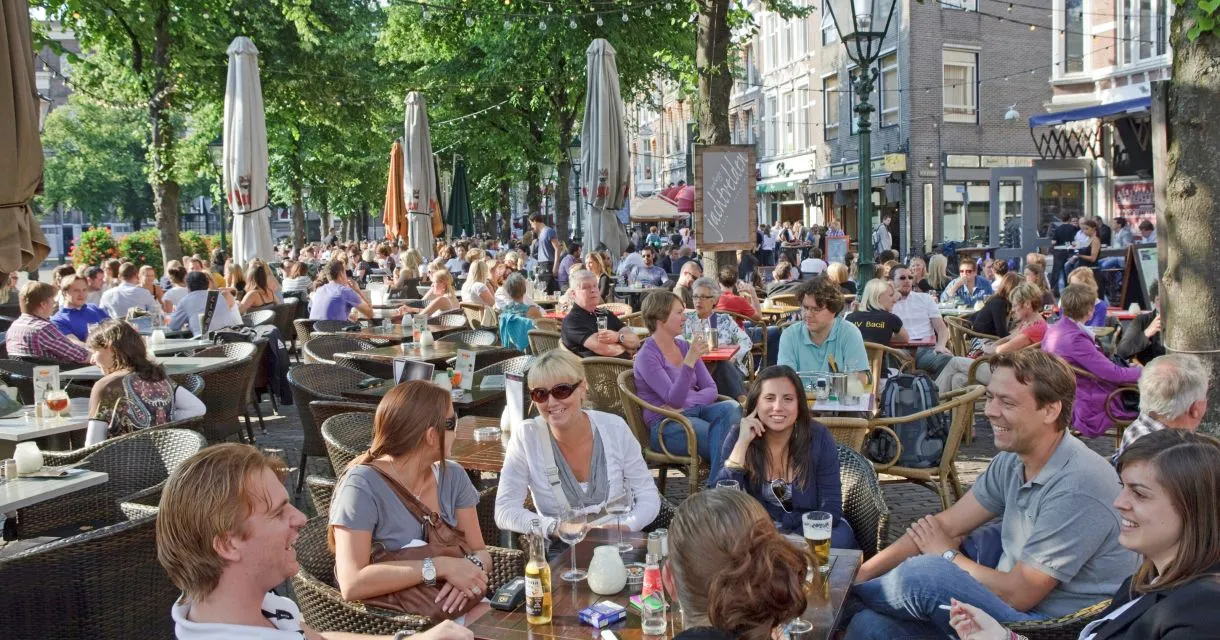 The image successfully captures a vibrant, photo-realistic scene of an outdoor cafe, with good color accuracy and clear focus on the main subjects in the foreground. The composition effectively conveys a lively atmosphere. However, the lighting is somewhat harsh, leading to strong contrasts and some areas of deep shadow or overexposure.