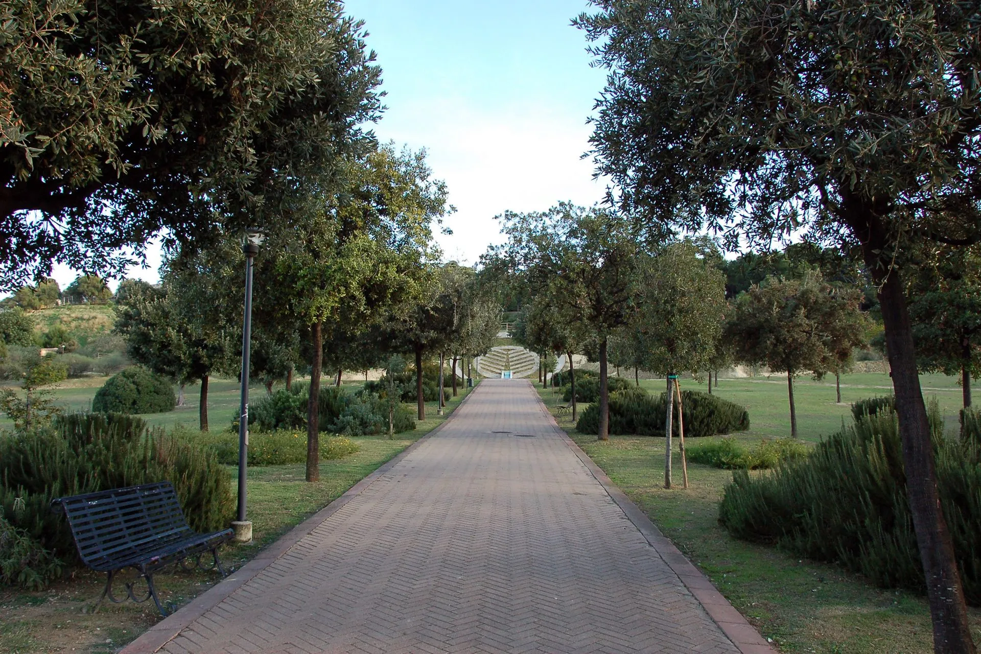 An editorial image showing a long, paved pathway made of reddish bricks, flanked by green grass, shrubs, and numerous trees. The path extends into the distance, leading towards a set of tiered steps. The sky above is clear and light blue. A park bench is partially visible on the left side in the foreground, and a lamppost stands nearby.