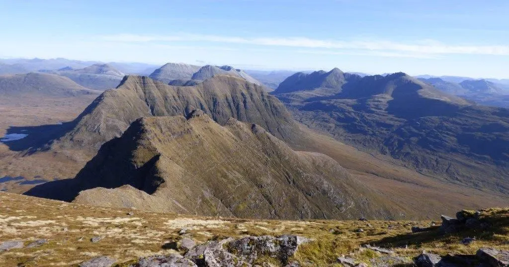 A wide-angle view of a vast mountainous landscape under a clear blue sky, featuring rugged peaks and valleys covered in sparse vegetation, with some rocks in the foreground.