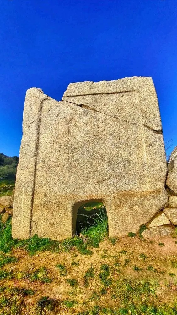 An editorial-style image featuring an ancient, weathered stone structure, possibly a megalithic monument or part of an old building, set against a bright blue sky with green grass in the foreground.