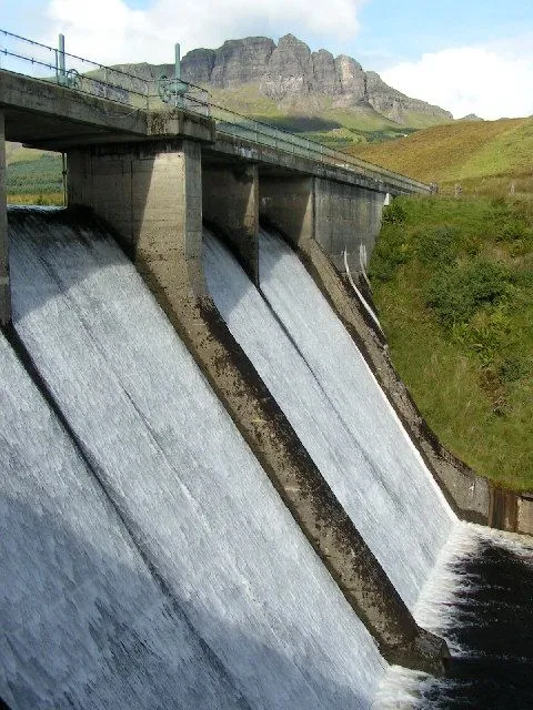 An image of a large concrete dam with water flowing over its spillways, set against a backdrop of green hills.