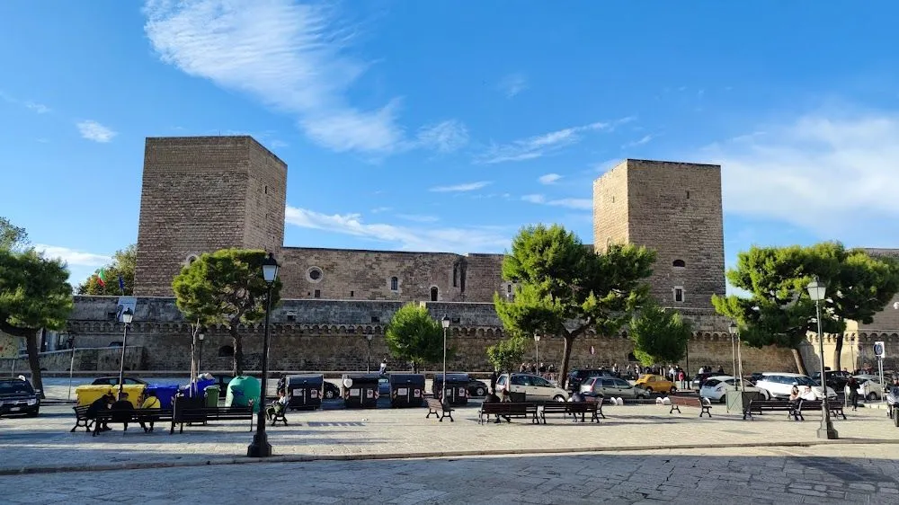 An exterior view of a large, old stone fortress or castle, seen from a public square with benches, lampposts, trees, and some parked vehicles under a clear blue sky.