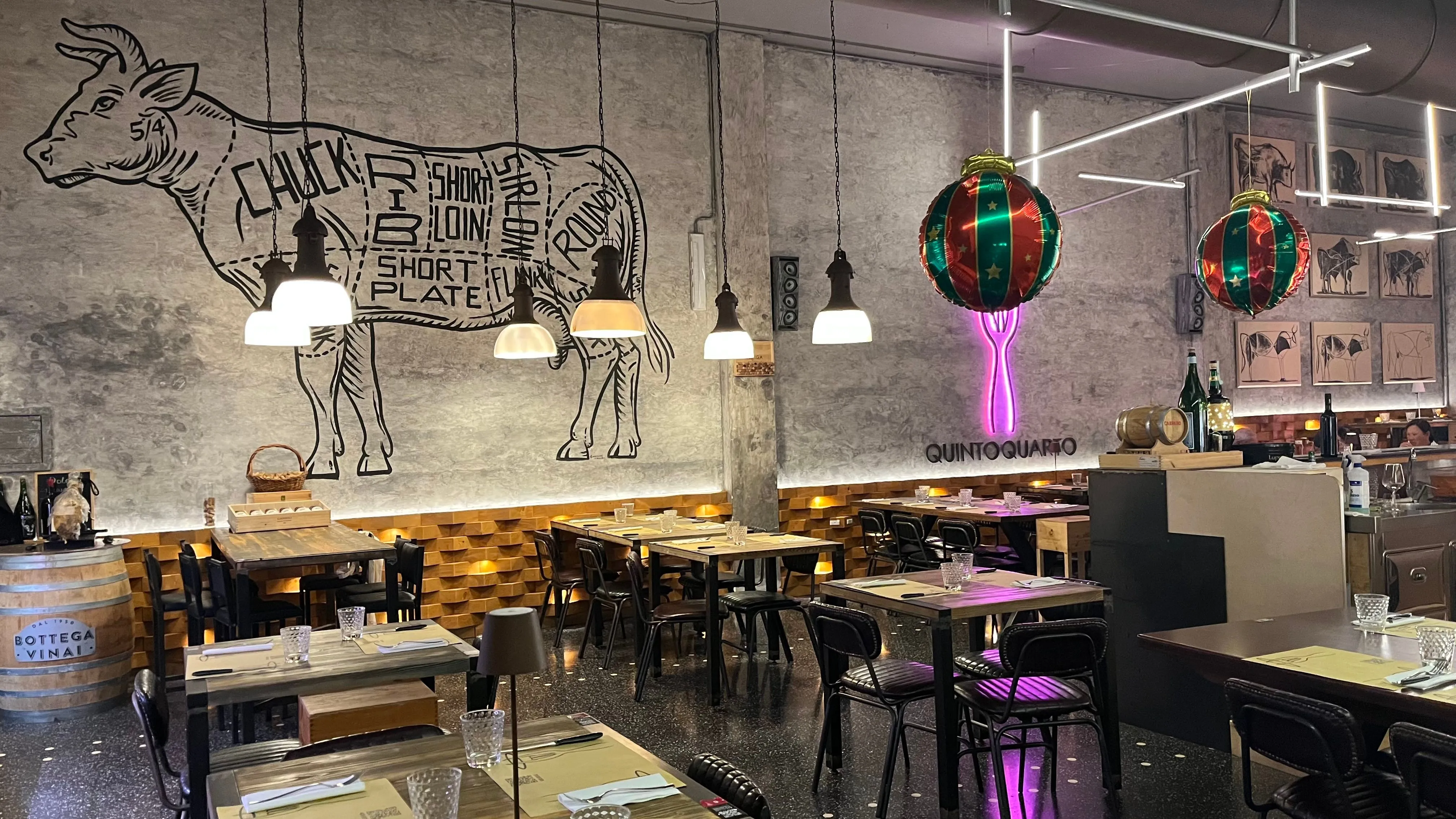 An interior view of a modern restaurant featuring a large wall mural depicting beef cuts. The space is furnished with simple wooden tables and black chairs, illuminated by hanging industrial-style lamps and subtle ambient lighting. A glowing pink neon fork sign and a large Christmas ornament add unique decorative elements.