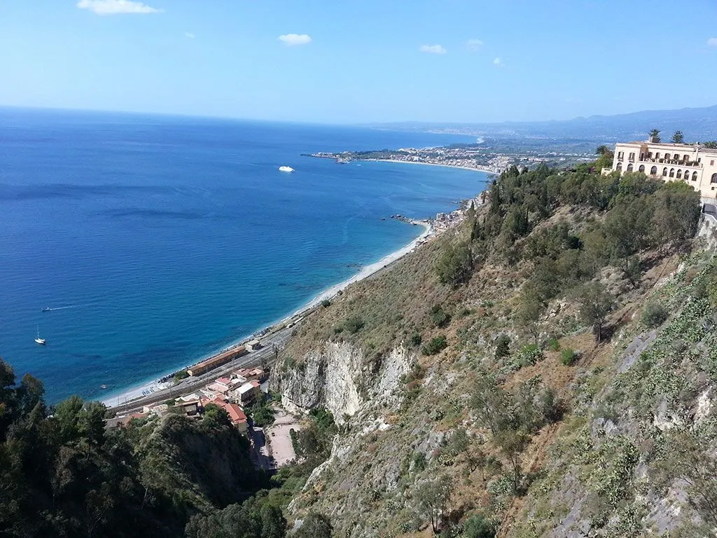 A high-angle panoramic view of a coastal landscape, featuring a vibrant blue sea, a long sandy and rocky shoreline, a small town nestled along the coast, and a steep, rugged mountain slope covered in vegetation in the foreground. A clear blue sky with minimal clouds completes the scene.