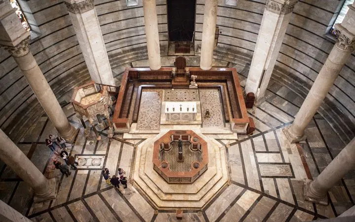 An aerial perspective captures the grand interior of a historic building, likely a baptistery or church. The image showcases intricate marble flooring with geometric patterns, a central octagonal baptismal font, and an ornate altar area. Large columns and detailed architectural elements define the space.