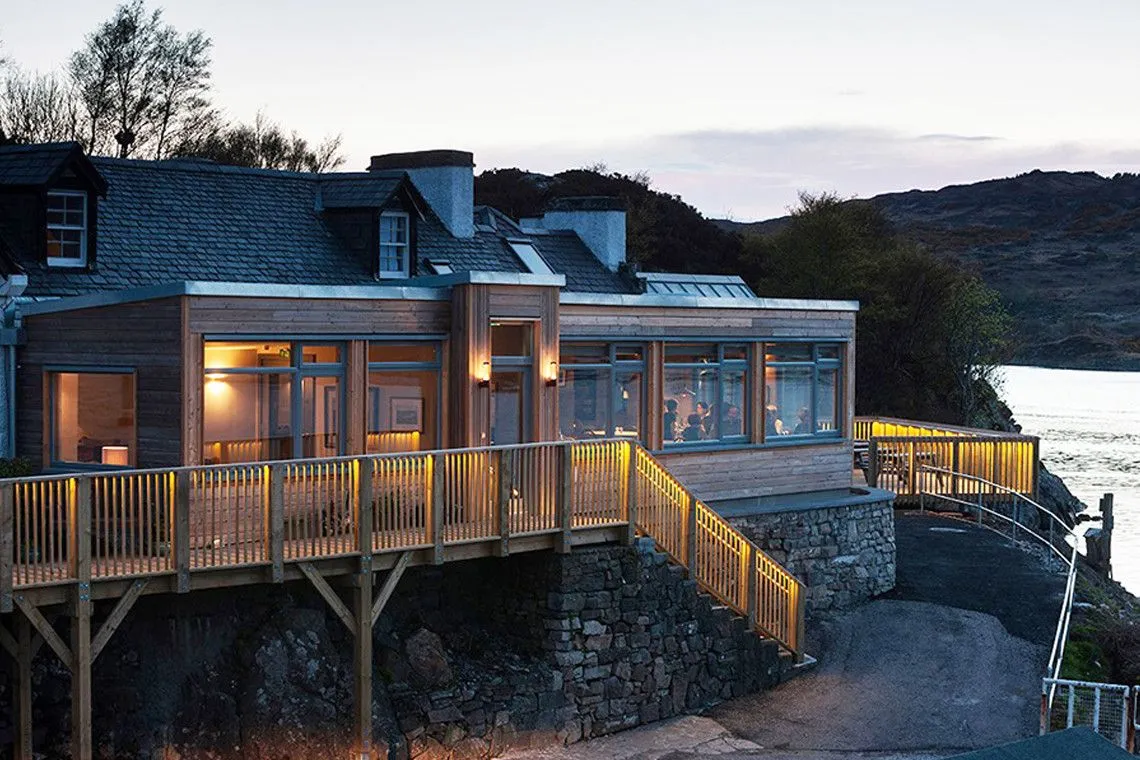 An inviting view of a modern building, likely a restaurant or hotel, at dusk. The warm illumination from inside and along the wooden deck creates a welcoming atmosphere.