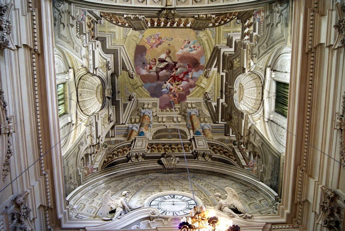 An upward view of an ornate church ceiling featuring a detailed fresco painting surrounded by intricate architectural elements, including decorative moldings, columns, and sculptures.