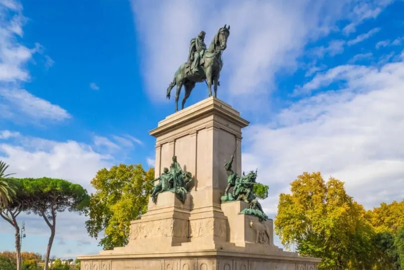A grand bronze equestrian statue on a stone pedestal stands prominently against a vibrant blue sky with scattered white clouds and verdant trees in the background.