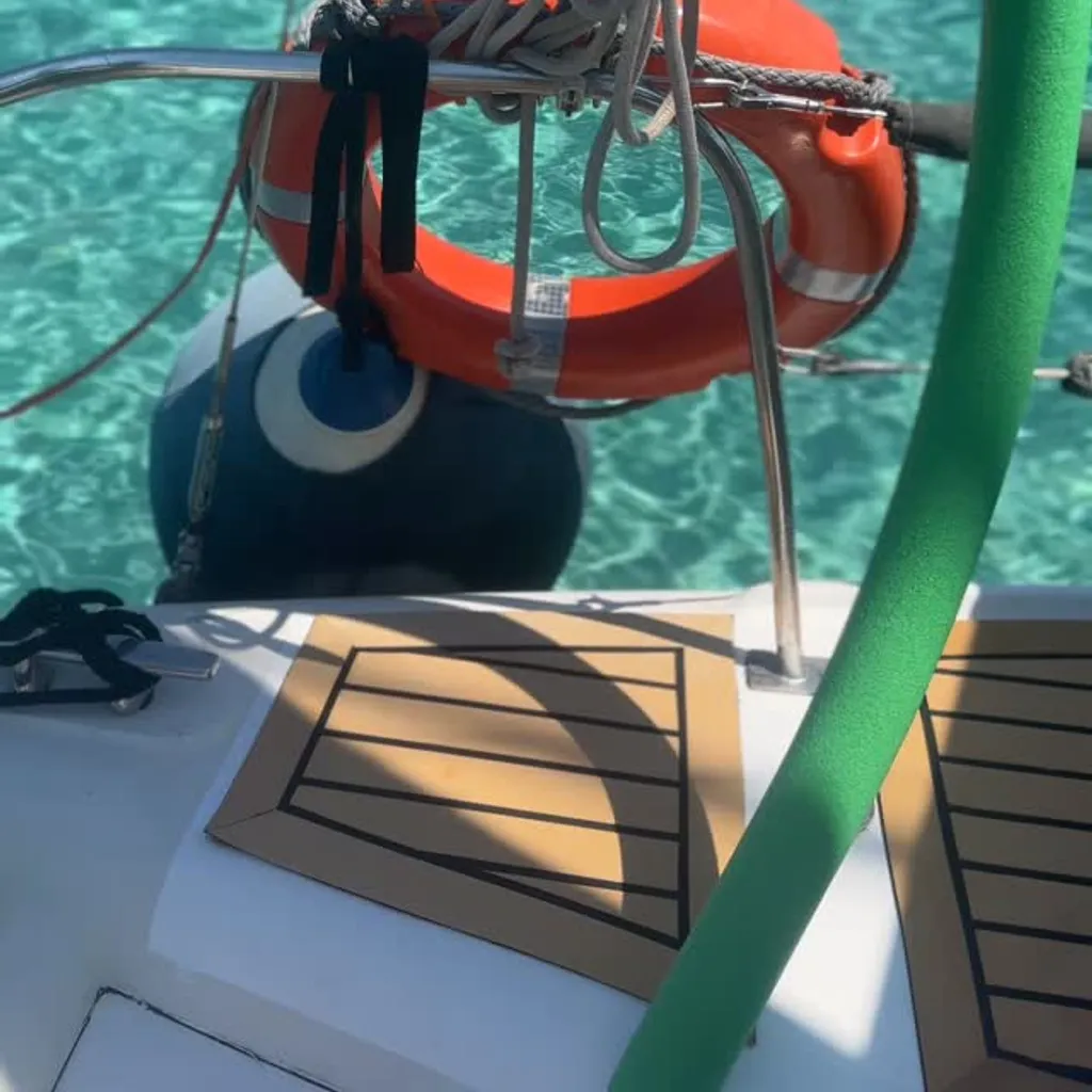 A close-up view from a boat, featuring parts of the vessel such as a metal railing, coiled ropes, an orange lifebuoy, and a blue fender. The background showcases crystal clear, turquoise water, indicating a serene marine environment. A prominent green foam-covered pole is visible on the right side of the frame.