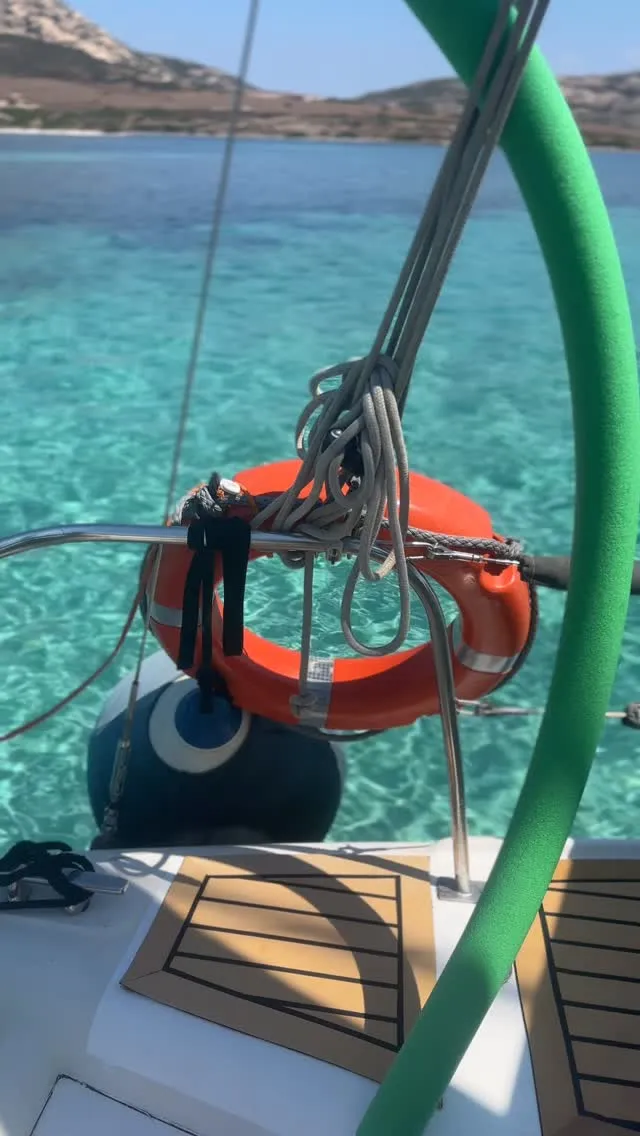 A close-up view from a boat, featuring parts of the vessel such as a metal railing, coiled ropes, an orange lifebuoy, and a blue fender. The background showcases crystal clear, turquoise water, indicating a serene marine environment. A prominent green foam-covered pole is visible on the right side of the frame.