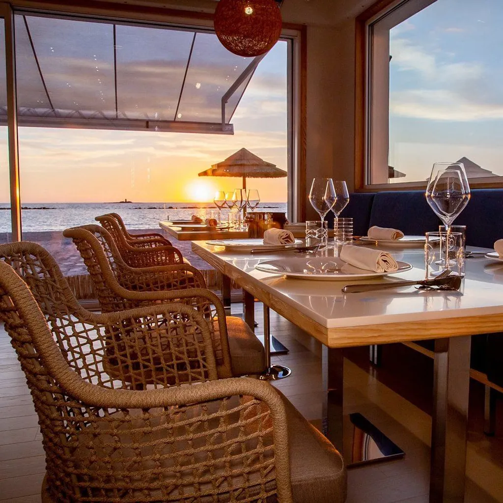A beautiful, well-composed image of a restaurant interior with a stunning sunset view over the ocean. The table is neatly set, and the scene evokes a peaceful, romantic atmosphere.