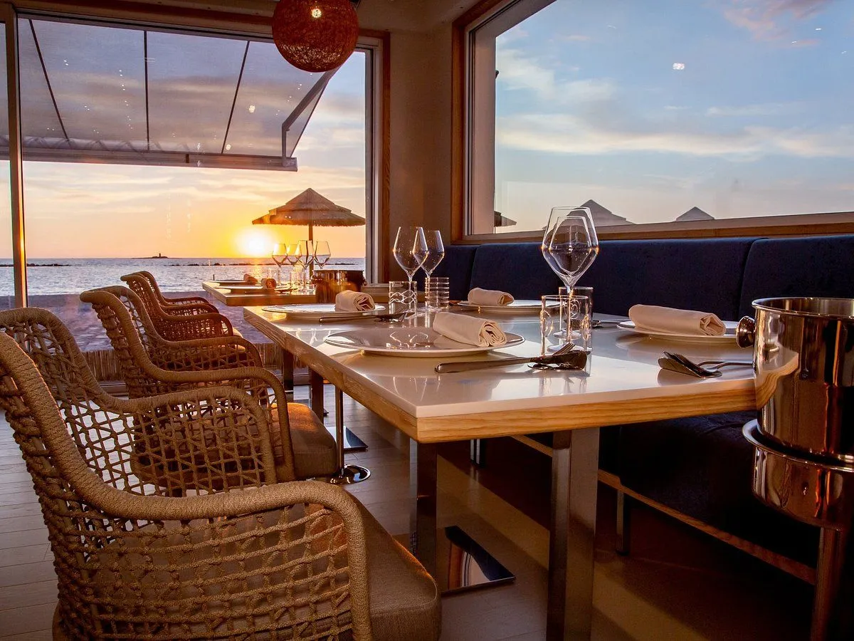 A beautiful, well-composed image of a restaurant interior with a stunning sunset view over the ocean. The table is neatly set, and the scene evokes a peaceful, romantic atmosphere.