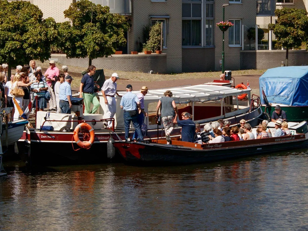 A lively scene depicting people boarding and disembarking from two canal boats, with a residential building and trees in the background under natural daylight.