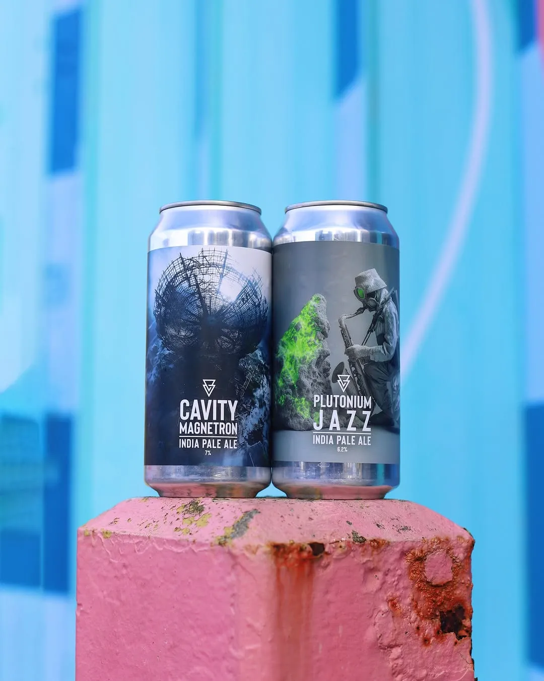 A high-quality product photograph featuring two distinctively designed beer cans standing on a textured pink block, set against a soft, blurred blue background.