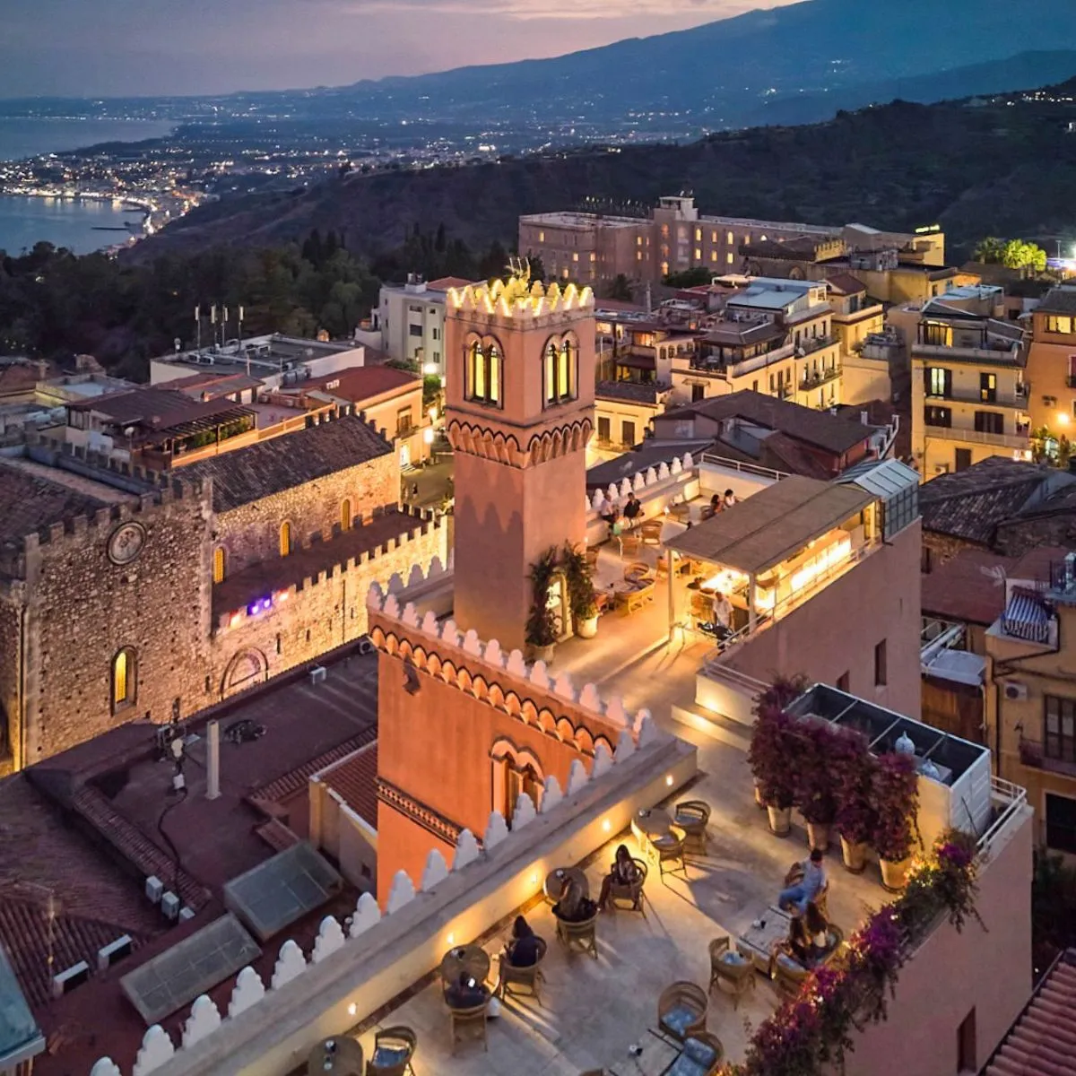 An aerial view of a charming coastal town at dusk or night, featuring illuminated historic buildings and a vibrant cityscape along the coastline, with distant mountains.