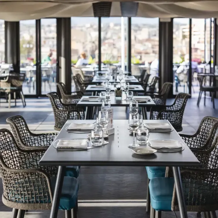 An inviting and well-composed image of a modern restaurant interior, featuring neatly arranged tables and chairs, bathed in natural light from large windows that offer a panoramic view. The setting appears ready for service, with all place settings meticulously laid out.