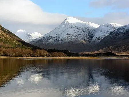 A stunning winter landscape featuring snow-capped mountains reflecting in a calm lake under a bright, partly cloudy sky. The foreground shows the still water of the lake, followed by a strip of trees and then the imposing snow-covered peaks.