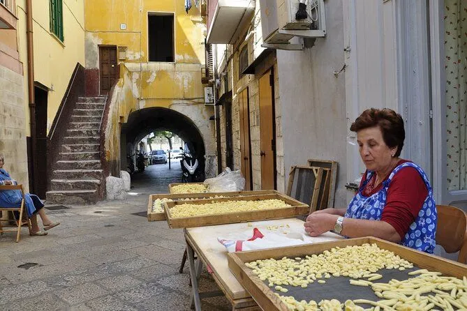The image captures a woman making traditional pasta, likely orecchiette, on a table set up in a narrow, rustic street, with an ancient archway leading to a continuation of the street in the background. Several trays of pasta are visible on and around the table.