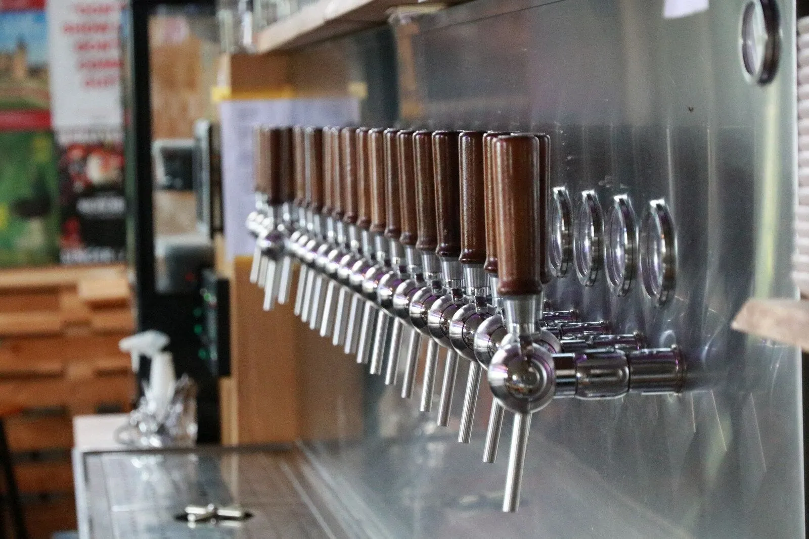The image features a neatly arranged row of beer taps with wooden handles, showcasing a bar or brewery setup. The foreground taps are sharp and well-lit, with a good depth of field that blurs the background slightly. The metallic and wooden textures are clearly visible, contributing to a realistic and inviting portrayal of the serving area.