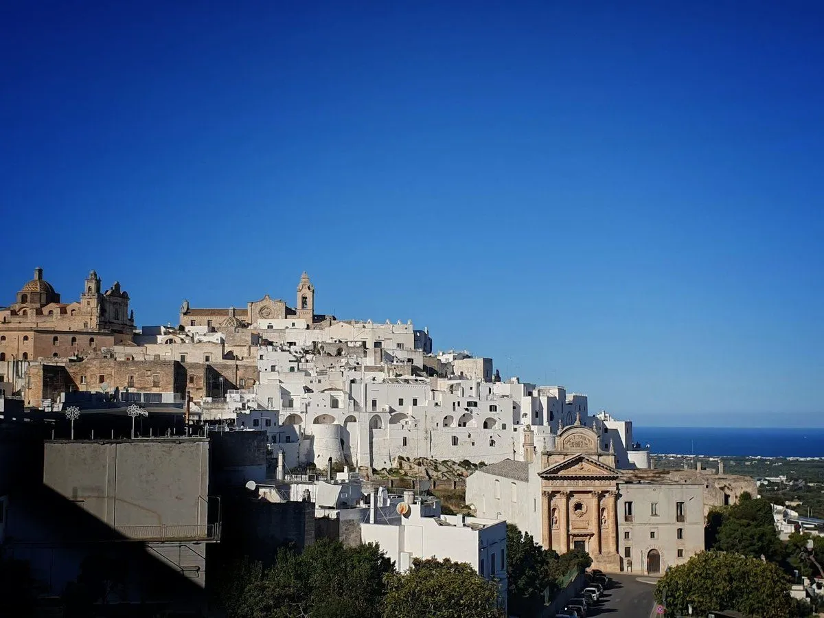 An editorial style image featuring a hilltop town with whitewashed buildings under a clear blue sky, with the sea visible in the distance. The architecture is prominent against the bright natural light.