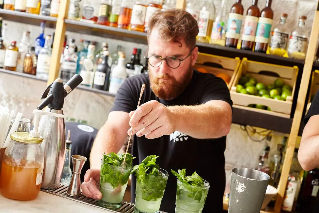 An image of a bearded male bartender wearing glasses, stirring drinks that appear to be mojitos behind a bar. The foreground clearly shows the drinks and his hands, while the background features liquor bottles on shelves, indicating a bar environment.
