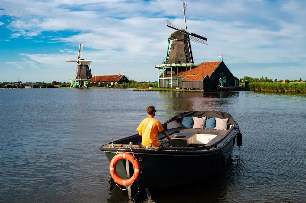 An editorial style image depicting a person in a small boat navigating a body of water, with traditional Dutch windmills and houses dotting the landscape in the background under a bright, partly cloudy sky.