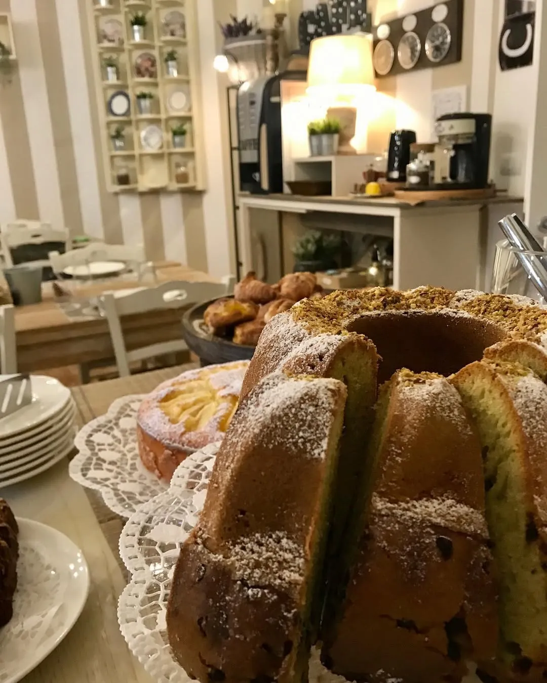 A close-up shot of a sliced bundt cake dusted with powdered sugar and topped with nuts, likely pistachios, placed on a lace doily. Another smaller cake is partially visible behind it. The background shows a soft-focused interior of what appears to be a cafe or buffet setting with tables, chairs, and kitchen equipment.