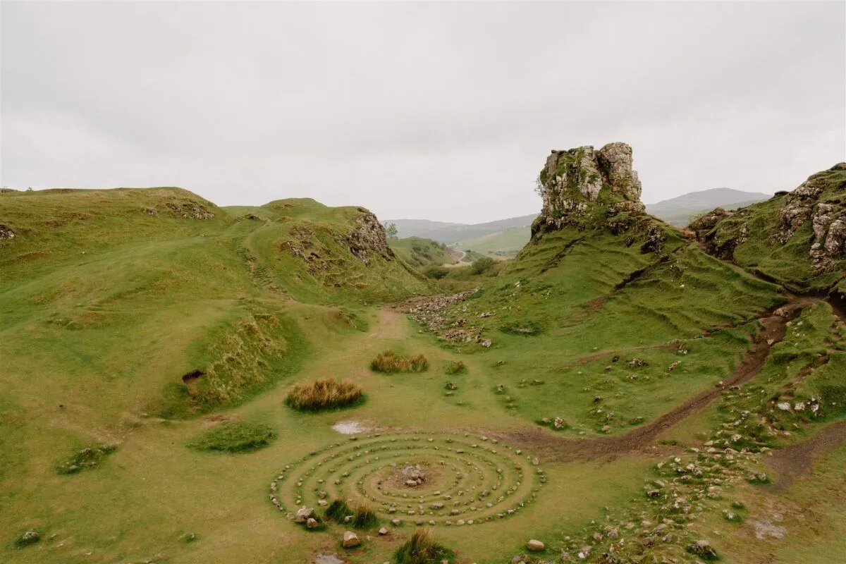 A wide shot of a green, rolling landscape under an overcast sky, featuring a prominent rock formation and a spiral pattern made of stones in the foreground.