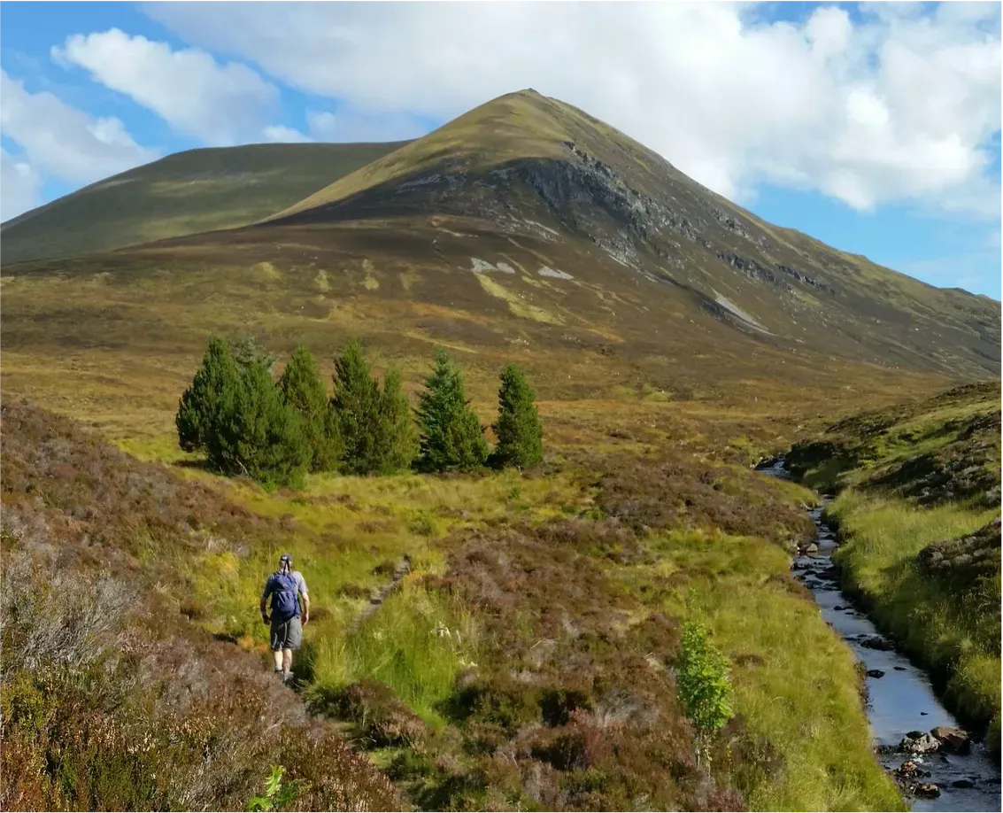 A well-composed landscape photograph featuring a hiker traversing a path through a scenic mountainous and moorland environment. The image is well-lit by natural daylight, showcasing the greens and browns of the terrain under a partly cloudy sky.