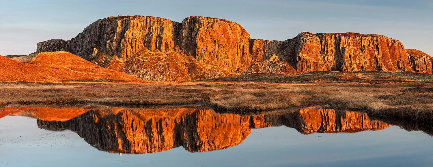 An exceptionally well-composed landscape photograph capturing a large, reddish-orange rock formation perfectly reflected in a calm body of water. The image benefits from dramatic golden hour lighting, which enhances the textures and colors of the rock. The clarity and sharpness throughout the image, coupled with natural and vibrant colors, contribute to its highly photo-realistic quality.
