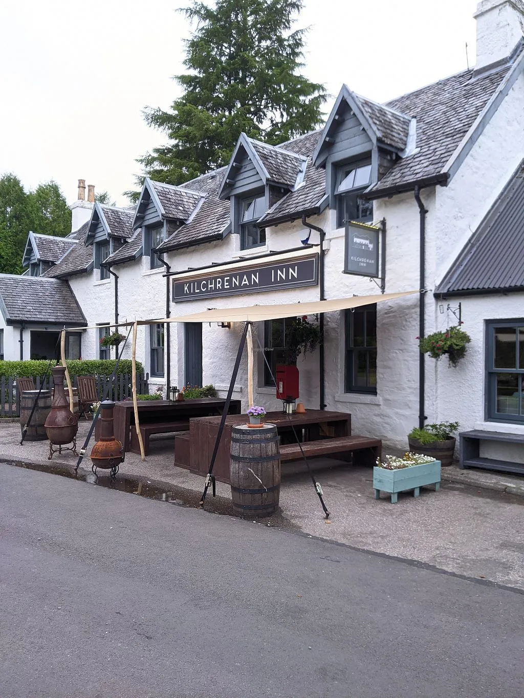 An exterior view of the Kilchrenan Inn, a white building with grey roof dormers, featuring an outdoor seating area with a beige sun tarp, wooden tables, and barrels.