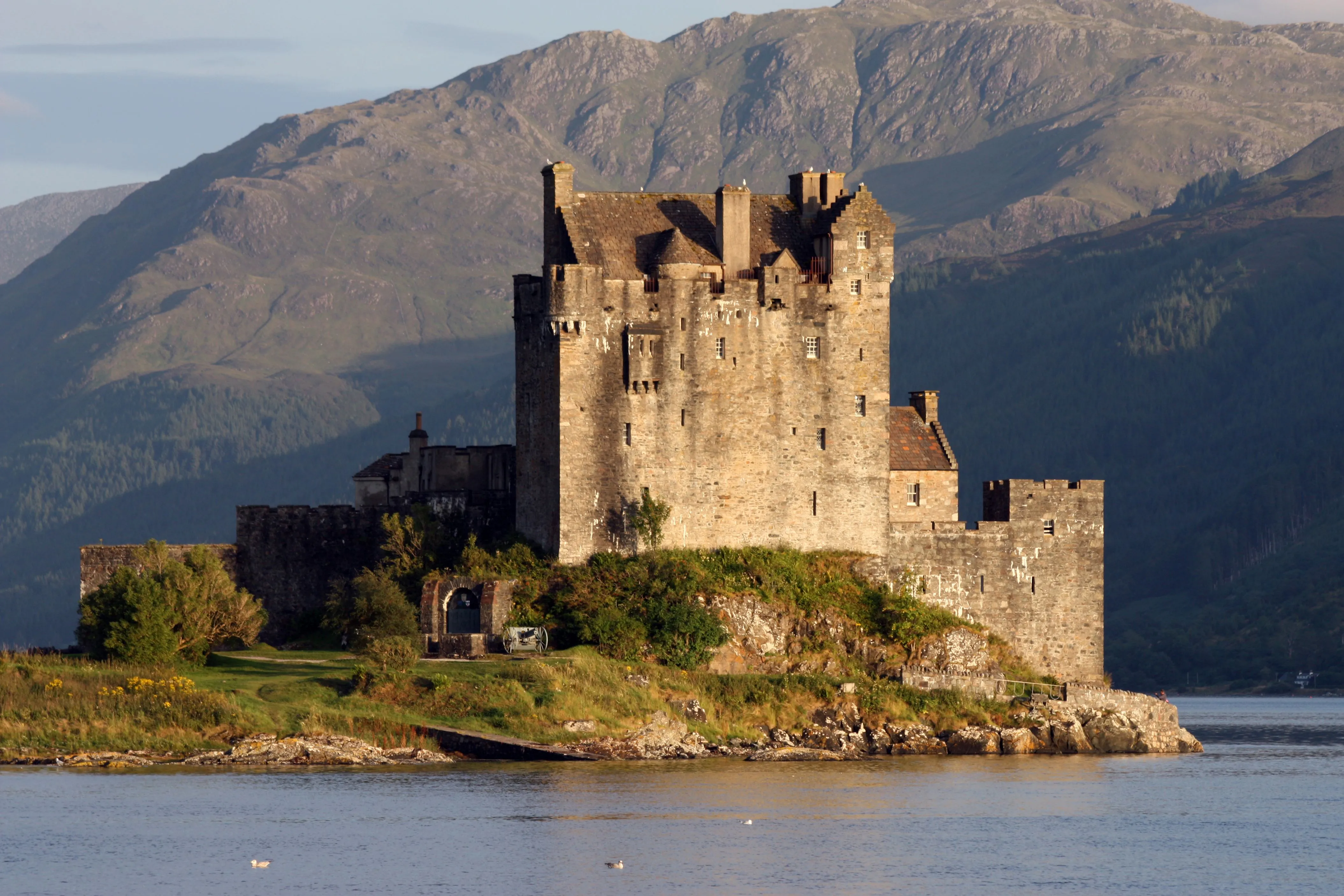A striking photograph of Eilean Donan Castle, majestically situated on a small island at the confluence of three sea lochs, with mountains rising in the background and calm water in the foreground.