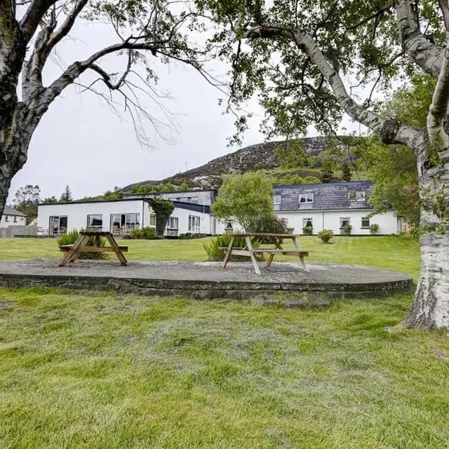 A photo of a white building with dark roofs, nestled in a green landscape with two picnic benches in the foreground. Large trees with light-colored bark frame the image. The sky is overcast, and a hill rises in the background.
