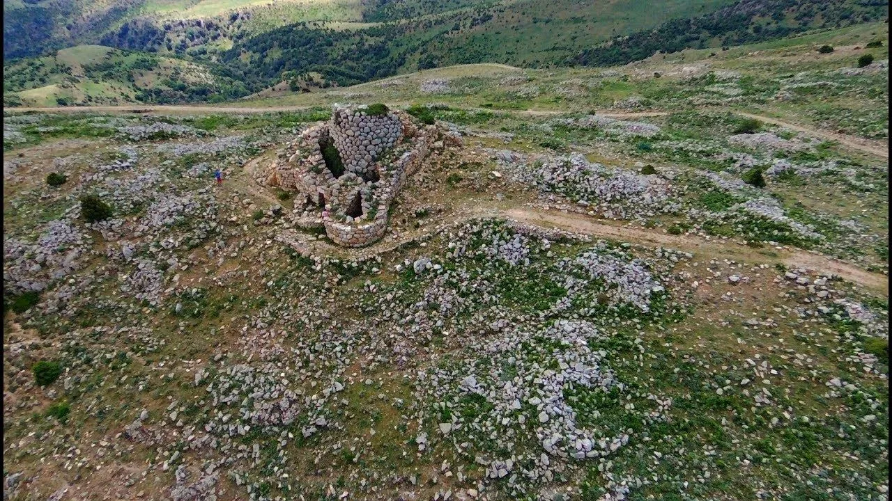 An aerial view of an ancient stone structure, possibly a nuraghe or similar ruin, nestled on a rocky, grassy hillside. The surrounding landscape consists of undulating green hills under what appears to be an overcast sky. Small figures of people are visible near the structure, providing a sense of scale.