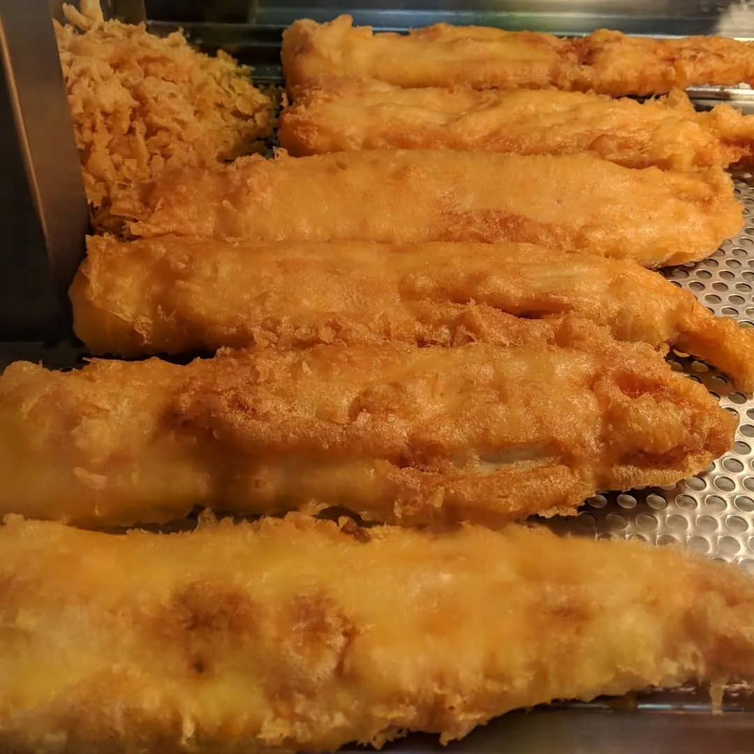 A close-up image showcasing several pieces of golden-brown fried fish, likely battered, resting on a metallic grate. The texture of the crispy batter is clearly visible, and the fish appears fresh and hot, suggesting it's in a food warmer or display.