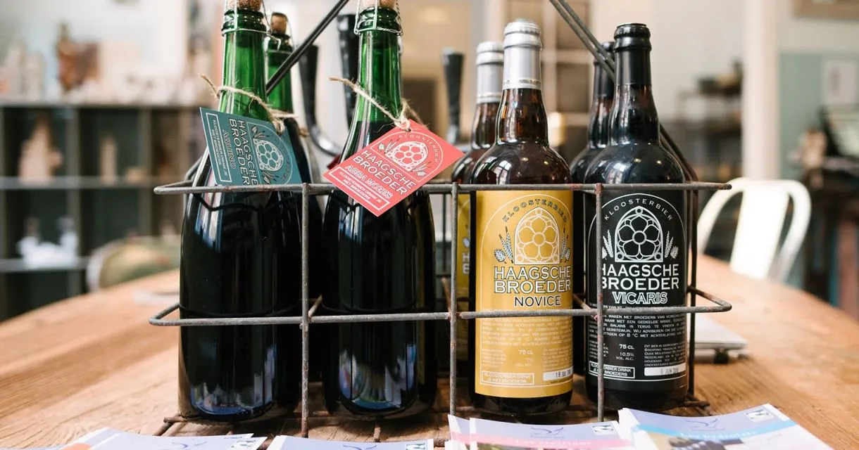 An editorial-style image featuring multiple bottles of 'Haagsche Broeder' beer, displayed within a metal rack. The foreground bottles, with their distinctive labels and corked tops, are in sharp focus, while the background is softly blurred.