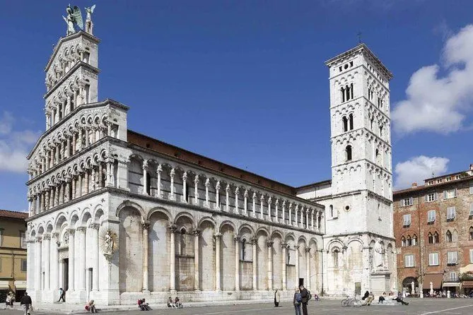 An impressive exterior view of a historic cathedral in Lucca, Italy, showcasing its intricate facade and a prominent bell tower under a clear blue sky. The image captures the grandeur of the architectural landmark.