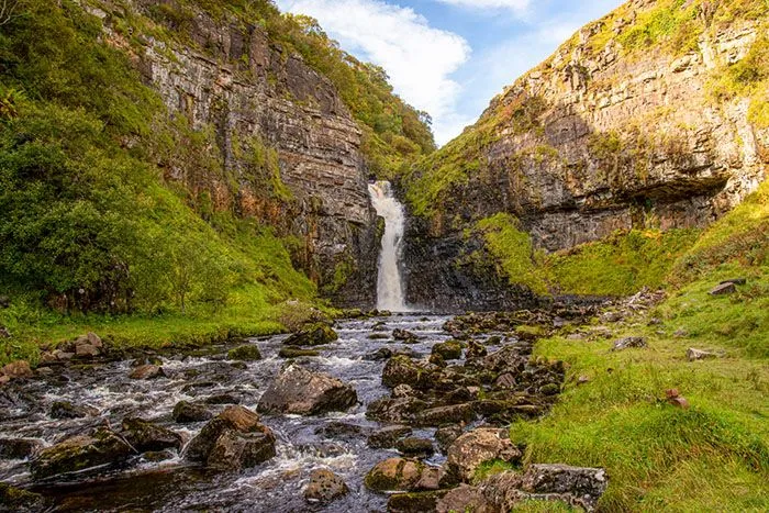 A stunning landscape photograph featuring a powerful waterfall cascading between two verdant, rocky cliffs, with a river flowing over moss-covered rocks in the foreground under a partly cloudy sky.