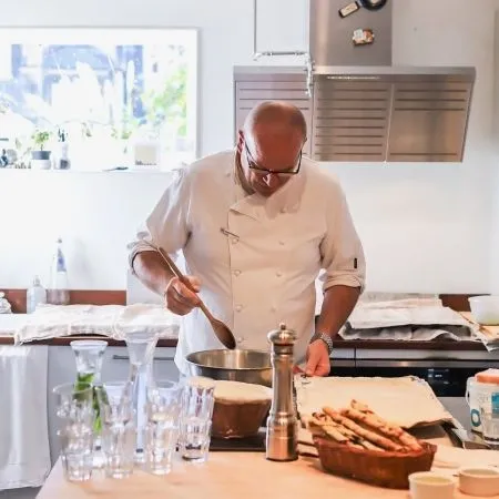 The image features a male chef actively cooking in a well-lit kitchen. The composition is balanced, focusing on the chef and his task. Key elements like the chef, the spoon, and the bowl are sharp and in focus. The colors appear accurate, contributing to a photo-realistic quality. While generally well-lit, there is slight overexposure in the background window, and the counter includes various items, though not overtly cluttered for an editorial context.