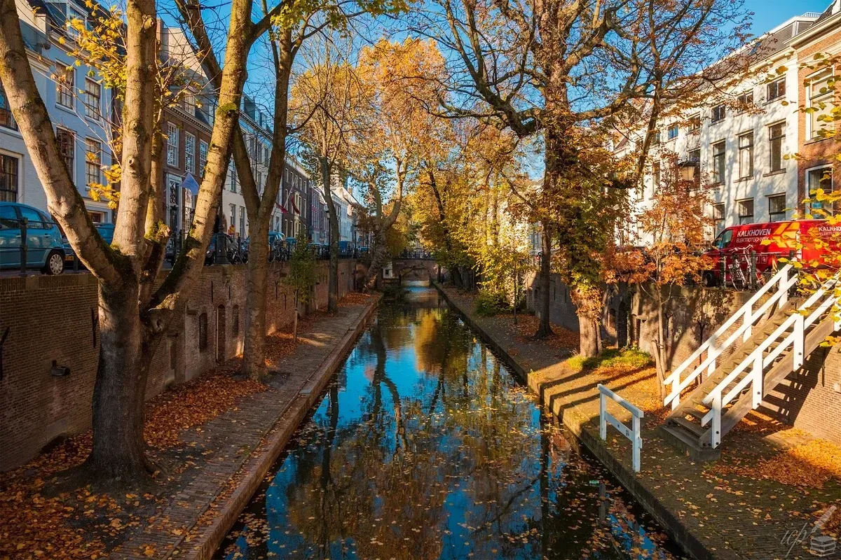An aesthetically pleasing image of a canal lined with buildings and trees in vibrant autumn colors. The reflections in the water are clear, enhancing the overall composition. The scene is well-lit and appears sharp with accurate, rich colors, making it highly photo-realistic.