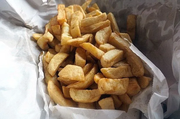 The image displays a generous serving of golden-brown chips (fries) wrapped in white paper. The chips appear freshly cooked and crispy, filling the frame. The lighting is even, highlighting the texture and color of the food. There are no unwanted elements present in the image.