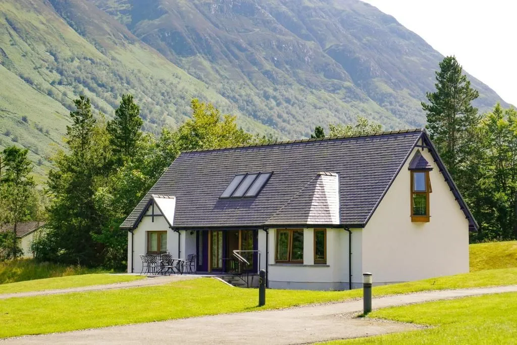 An exterior shot of a modern, white-walled house with a dark gray roof, featuring multiple windows and a patio area with outdoor furniture, set against a backdrop of lush green mountains under a bright sky.
