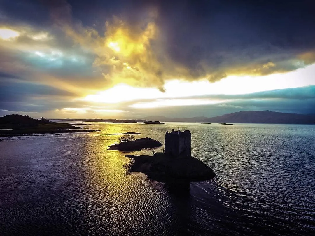 An atmospheric shot of a historic castle on a small island, silhouetted against a dramatic sunset or sunrise sky, with the sun's reflection shimmering on the surrounding water.