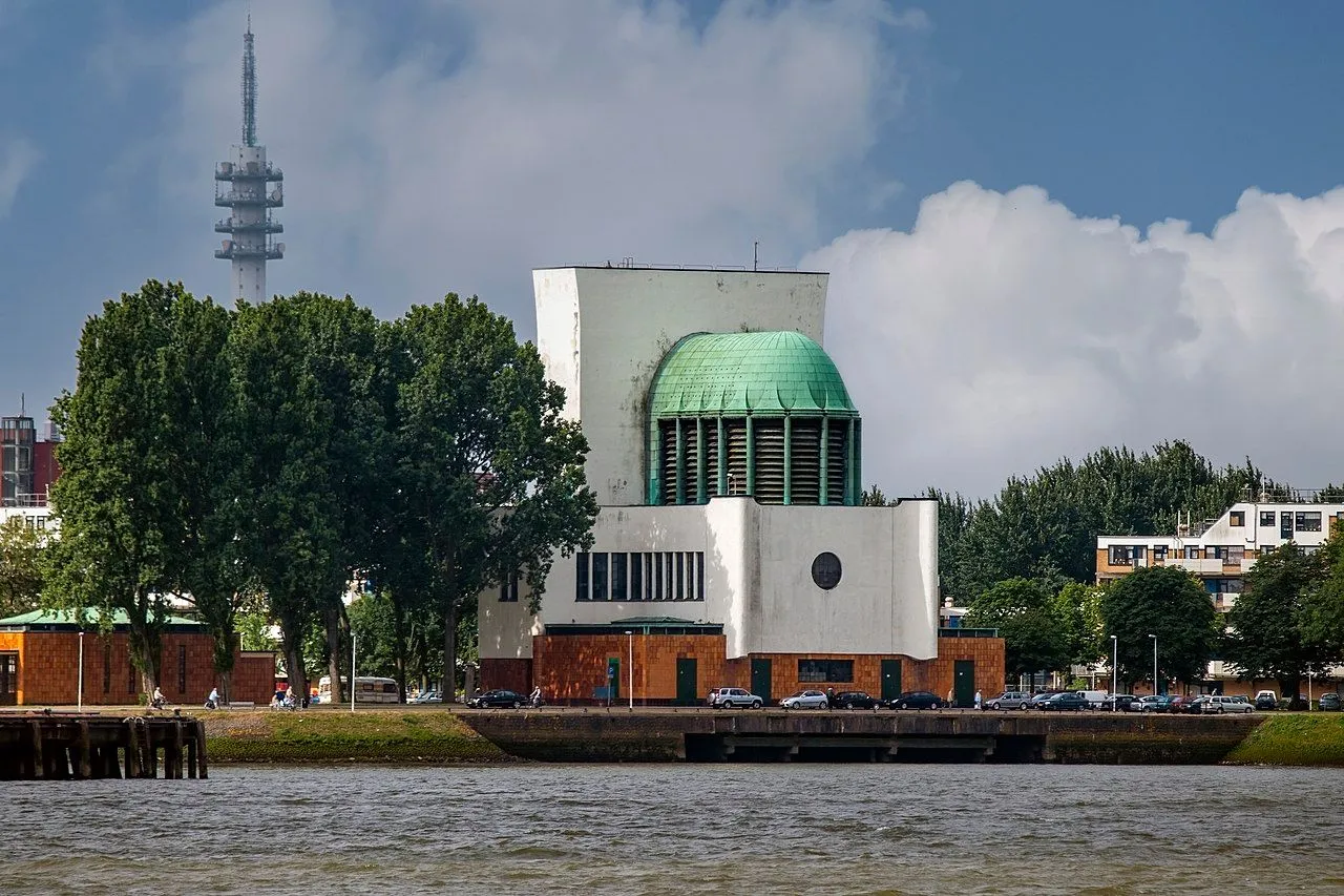 A well-composed and sharp photograph showcasing a distinct building with a prominent green dome, situated along a river. The image captures the architectural details clearly against a backdrop of trees and a distant telecommunications tower under a partly cloudy sky.