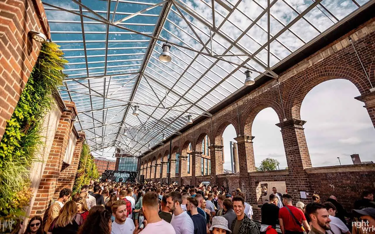 An editorial image capturing a large crowd gathered at an event or concert under a grand glass-roofed structure, featuring prominent brick arches. The scene is brightly lit by natural light, showing people enjoying the atmosphere.