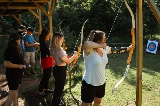 An outdoor photograph capturing a group of individuals, primarily women, engaged in archery. The main subject in the foreground is a woman in a white top and shorts, aiming a bow and arrow with focused concentration. Other participants are visible in the background, also holding bows, under a wooden shelter and surrounded by greenery.