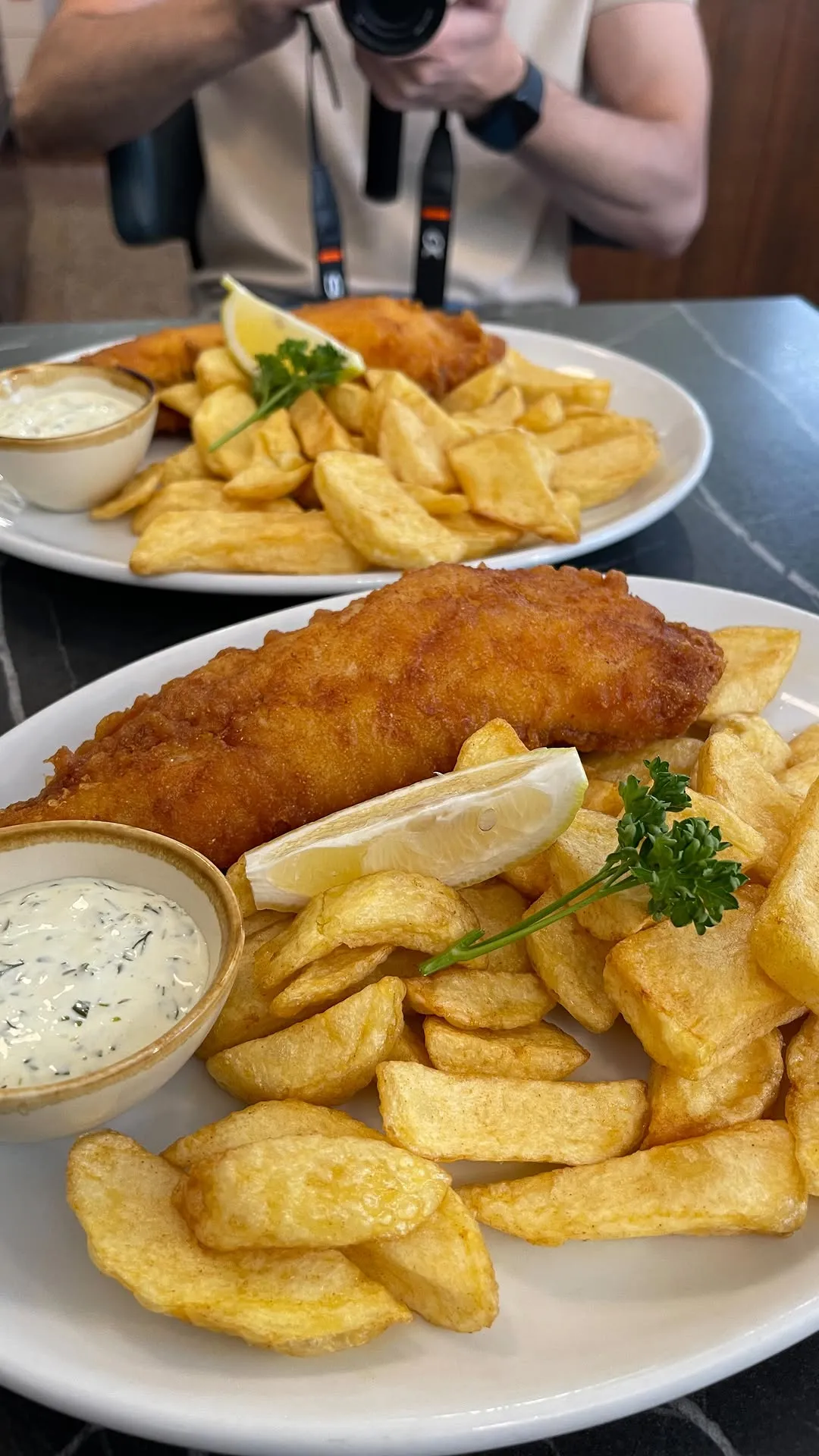 A close-up, photo-realistic image of two plates of fish and chips, with the foreground plate in sharp focus. The meal includes a golden-brown battered fish, chunky chips, a lemon wedge, and a side of tartare sauce, all presented on a white plate.