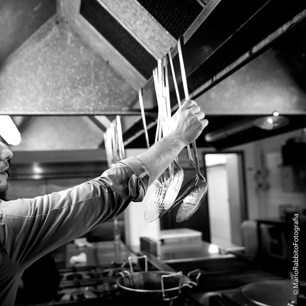 An editorial-style black and white image capturing a chef in a professional kitchen, reaching for cooking utensils. The image focuses on the action, highlighting the chef's hands and face, with kitchen equipment visible in the background.